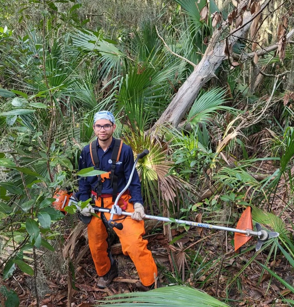 Jon Casado poses on downed tree holding vegetation cutter tool.