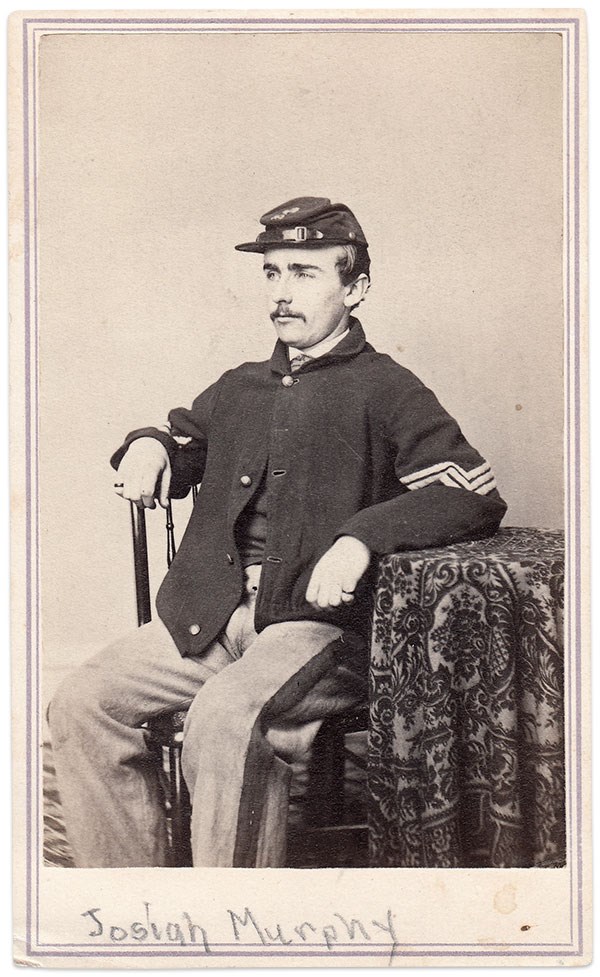 A Civil War solider in his early 20's sits posed next to a table.