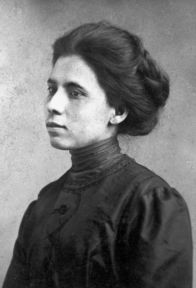 Portrait of a woman with dark hair pulled back from her face wearing a dark top with a collar that goes all the way up to her neck. Photograph is taken from the chest up with the woman turned 45 degrees away from the camera.