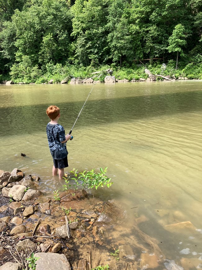 A young boy stands on a river bank holding a fishing pole with line in the water