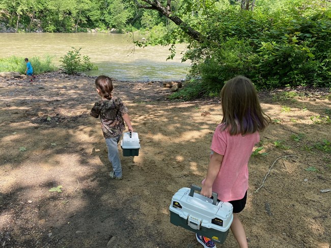 two children walk down to a riverbank holding plastic tackleboxes