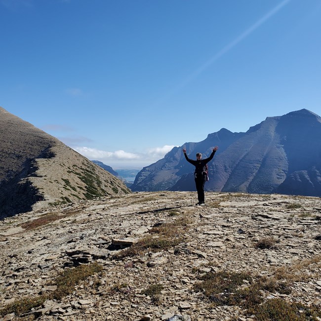 Woman in cold weather hiking clothing stands with outstretched arms overhead on top of a mountain peak.