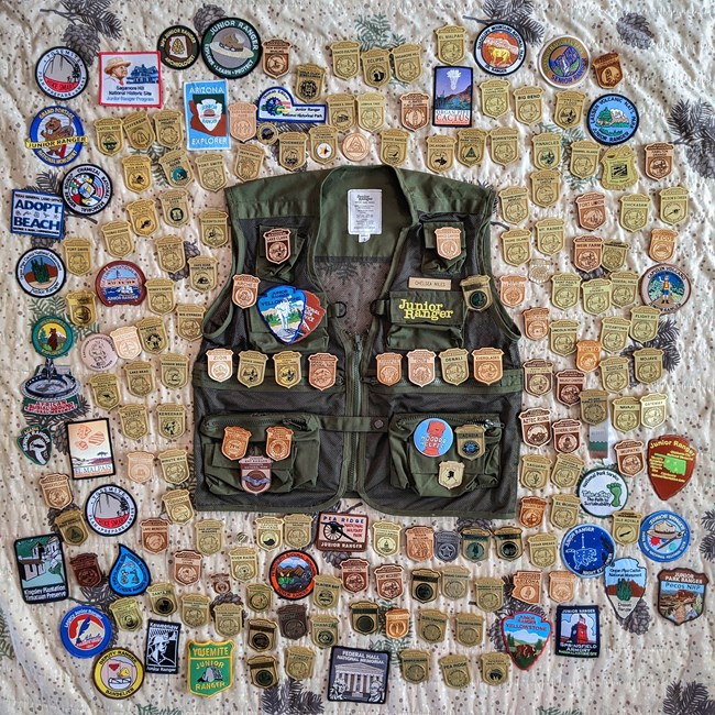 A junior ranger vest displays several junior ranger badges and patches, surrounded by approximately 100 junior ranger badges and park patches.