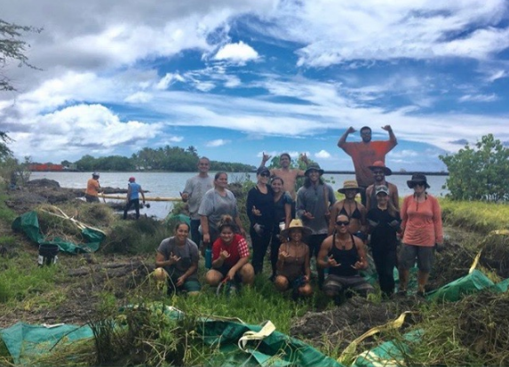 A group of muddy volunteers pose for a photo in front of Kaloko pond. Green bags of removed plants line the foreground.