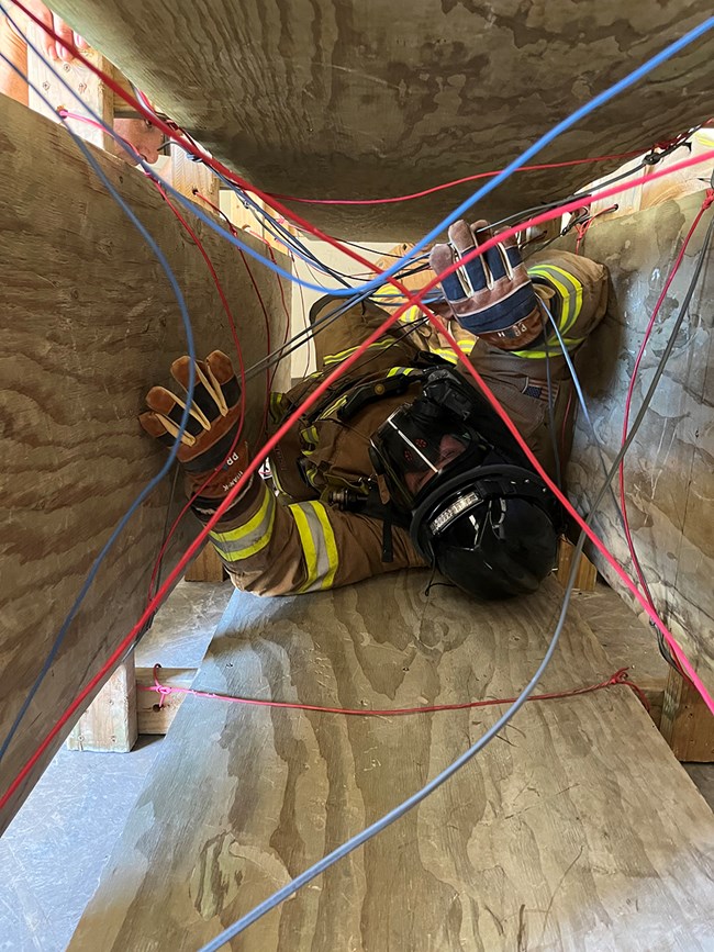A firefighter in full turnout gear is in a wooden box with wires obstructing the way forward.