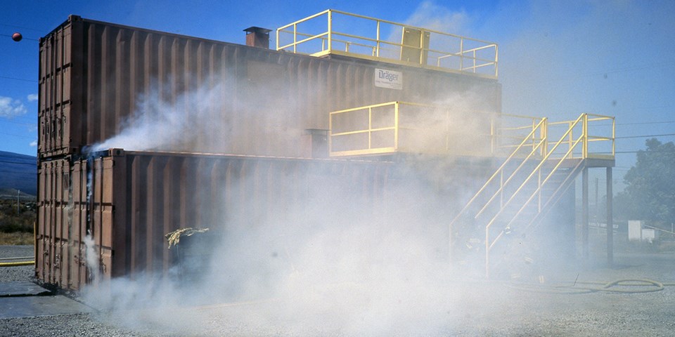 A structure made from shipping containers with stairs and railings added is obscured by smoke.