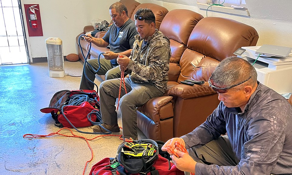 Three men sit on chairs in a room looking at ropes looped in their hands.