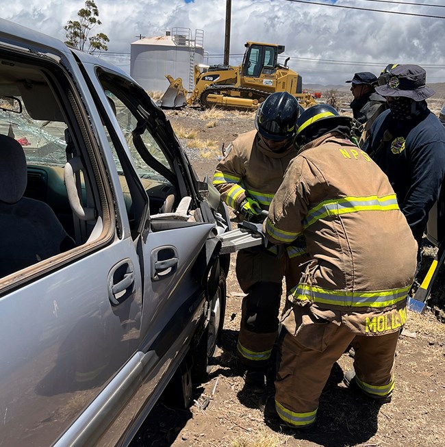 Two firefighters in turnout gear use a piece of equipment and bend a car door while two other people look on nearby.