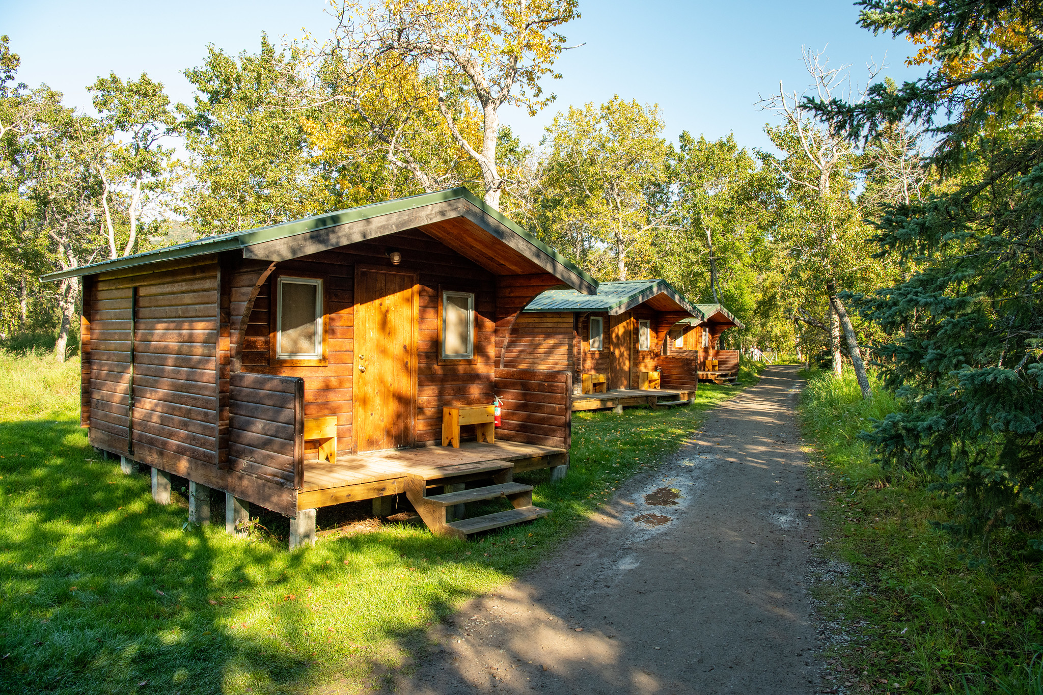 A row of three one-story wood cabins, each with a porch and overhanging eaves, at an angle to a dirt path.