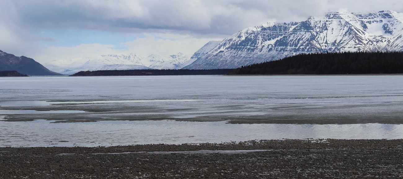 KATM View from shore to Naknek Lake A cover of snow highlights the contours of tall mountains, rising into clouds on the opposite shore of a lake.