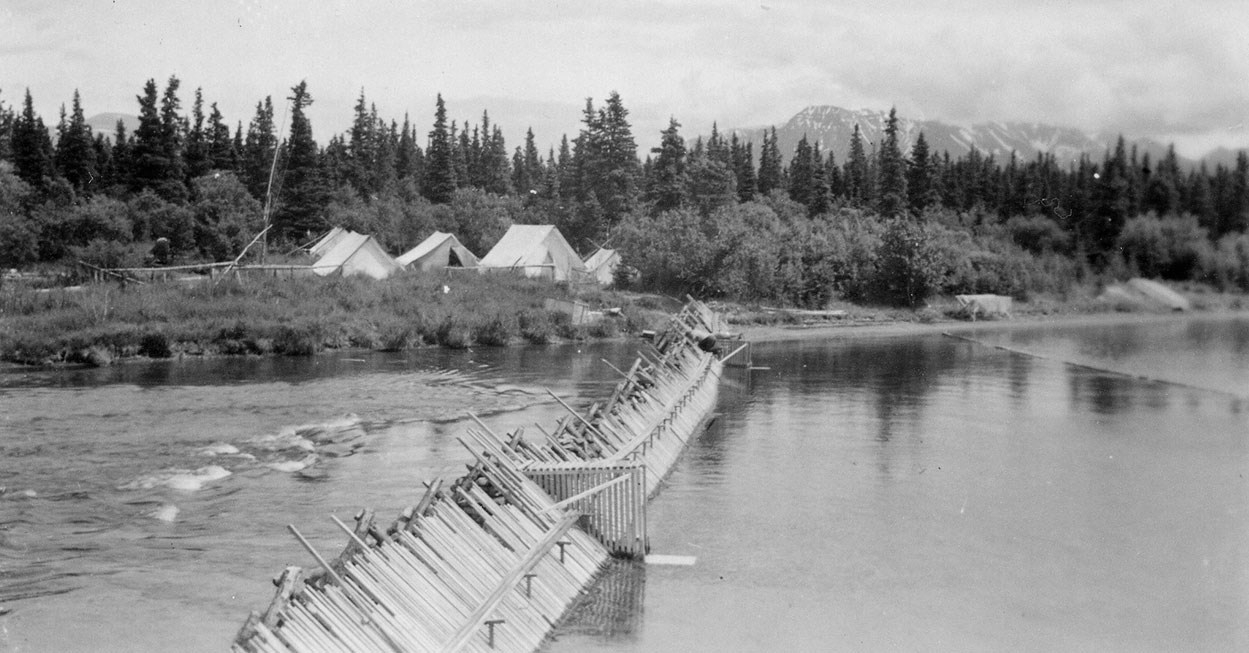 A cluster of tents stands on a shore across the mouth of a river, at the end of an angled wooden structure.