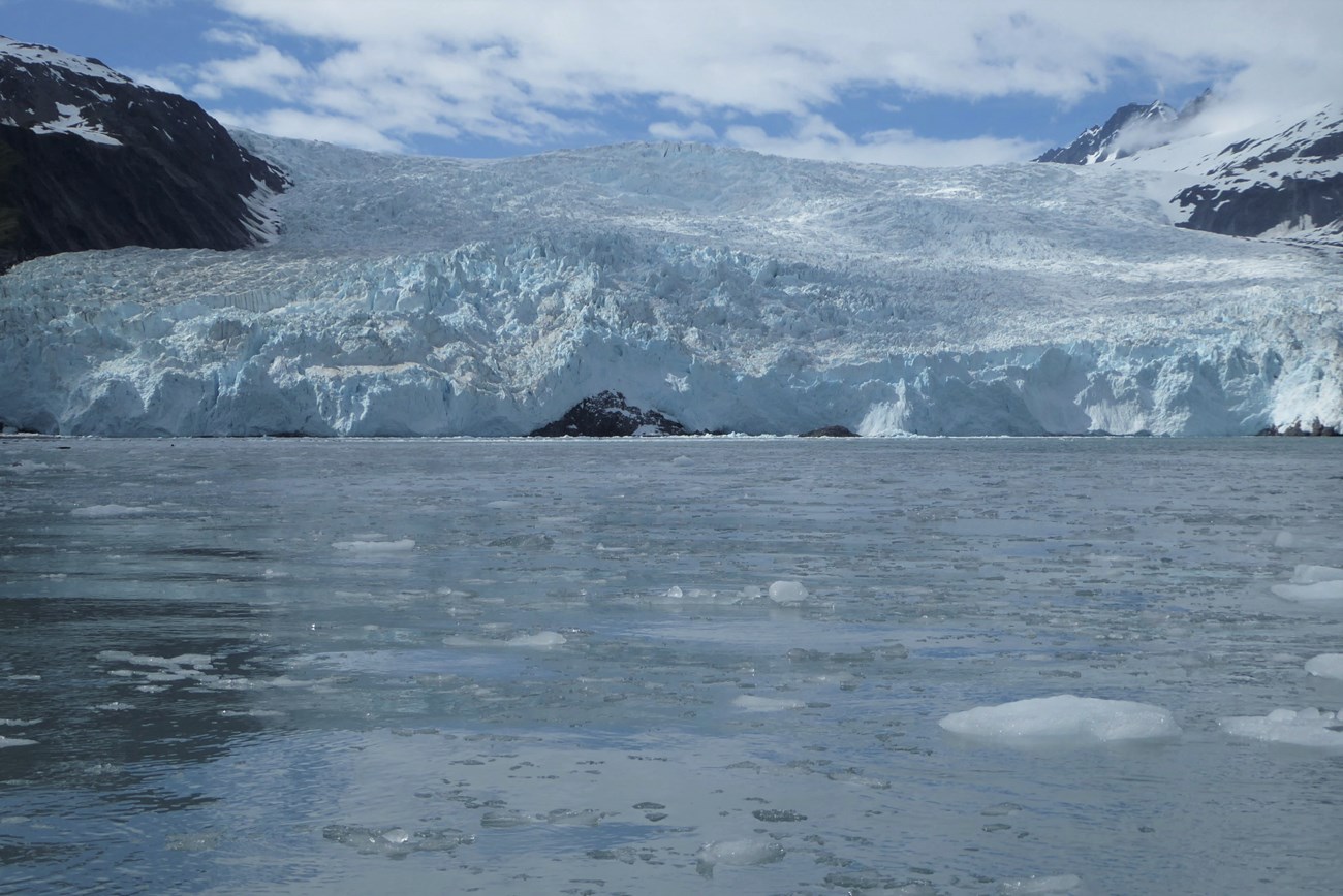 A massive glacier terminates in an icy bay. Dark rock masses appear at the base of the glacier before it reaches the water at some points.