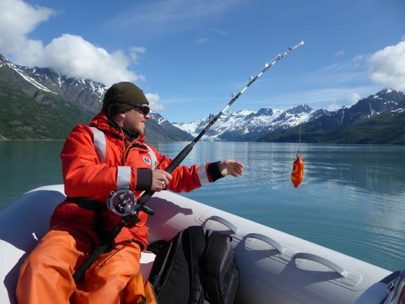 A researcher in a small skiff on the water reaches for a small orange CTD unit at the end of a fishing pole.