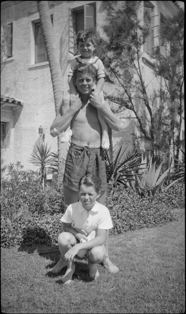 A black and white photo of a young Ted Kennedy sitting on John Kennedy's shoulders.  Bobby is seated below.