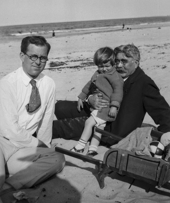 Three generations of Kennedys sit in the sand at Nantasket Beach.  There is a cart filled with beach supplies between them.  The ocean is seen in the background.