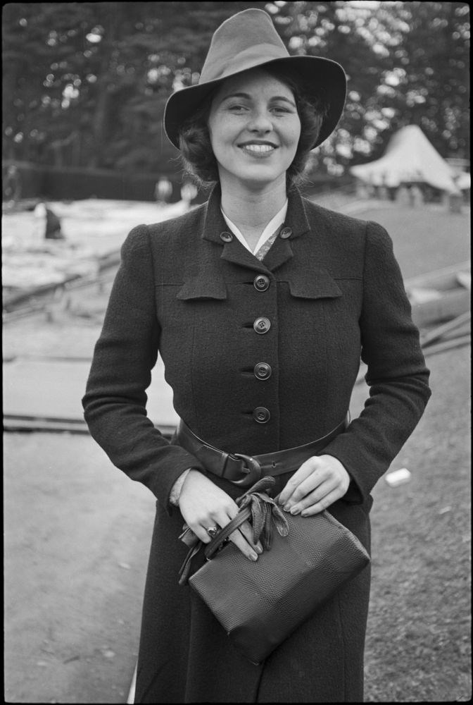 Rosemary stands holding a purse, wearing a large hat.