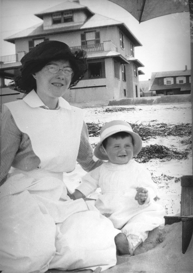 A nursemaid (left) sits with a toddler in the sand.  Behind them, sand, seaweed, and several cottages are visible.
