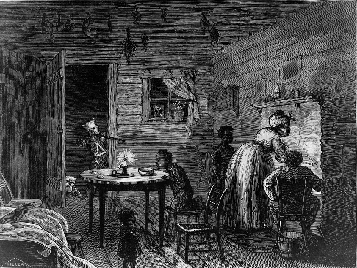 A white man wearing a white hood and holding a gun enters a house where African Americans are cooking at a fireplace.
