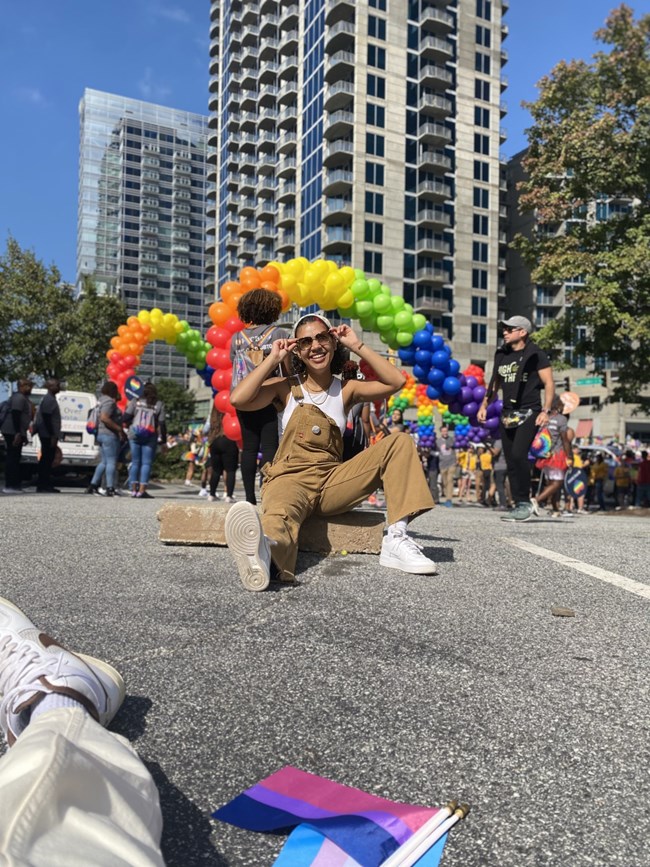 Kaia sits on the ground with rainbow balloons behind them at a pride festival.