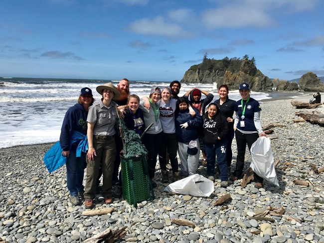 A group of teenagers and adults stand along a rocky beach holding bags of the littler they collected