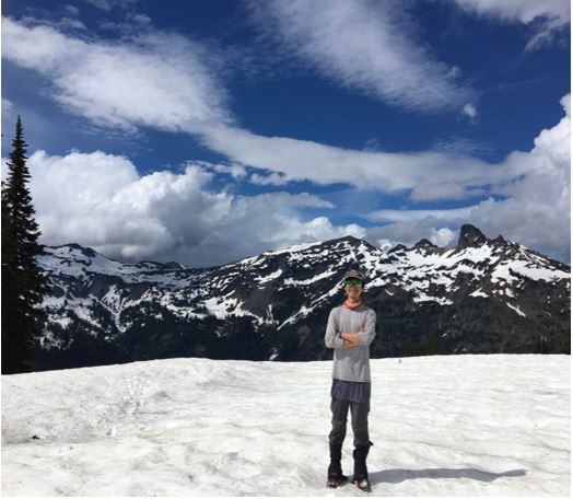 Young Asian male smiling with arms crossed, wearing a light gray shirt, dark gray pants, a gray hat, snow on the ground, mountains and trees in the background, with big fluffy white clouds and a blue sky