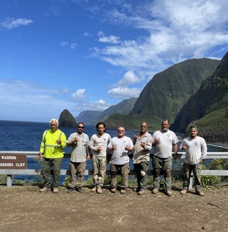 Seven men stand for picture, giving thumbs up. The steep, lush coastline of Molokai stretches into the distance behind them.