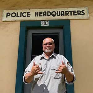 Kaiama is a 2022 winner of a regional Award for Cultural Resources Kaiama smiles in NPS uniform and holds up two hands in the “hang loose” sign, both pinkies and thumbs extended with the rest of his fingers folded down.