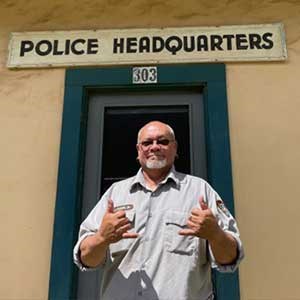 Kaiama smiles in NPS uniform and holds up two hands in the “hang loose” sign, both pinkies and thumbs extended with the rest of his fingers folded down.