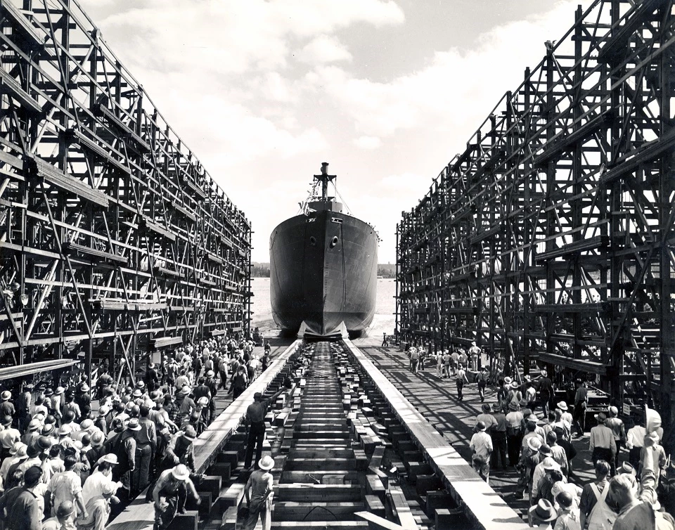 Kaiser Shipyards, Launching Ship - FOVA 219207 A black and white photo of a ship launching into the Columbia River. A crowd of workers stand with their backs to the camera, watching the ship slide into the water.