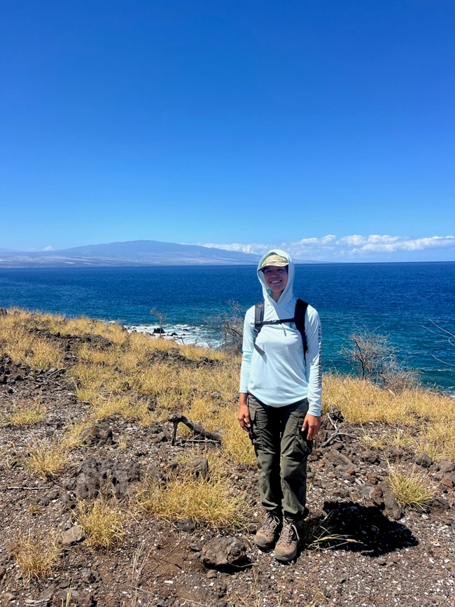 Karyssa poses for a photograph on level, rocky foreground with a view of the blue ocean in the background.