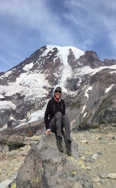Kai gets to experience a marmot’s lifestyle during a hike up Rainier Young Asian male smiling while sitting on a gray rock. Mountains with snow on them are behind him, along with a gray-ish blue sky. He is wearing a red shirt, a gray hoodie, black shirt, gray pants, and dark colored boots.