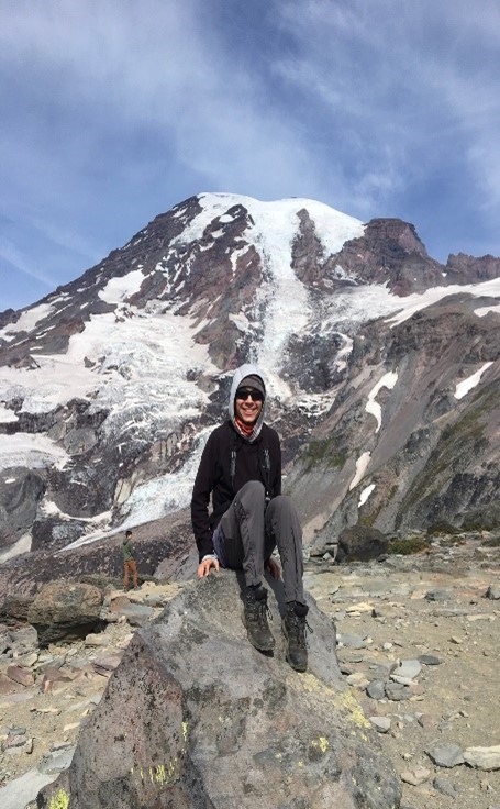 Young Asian male smiling while sitting on a gray rock. Mountains with snow on them are behind him, along with a gray-ish blue sky. He is wearing a red shirt, a gray hoodie, black shirt, gray pants, and dark colored boots.