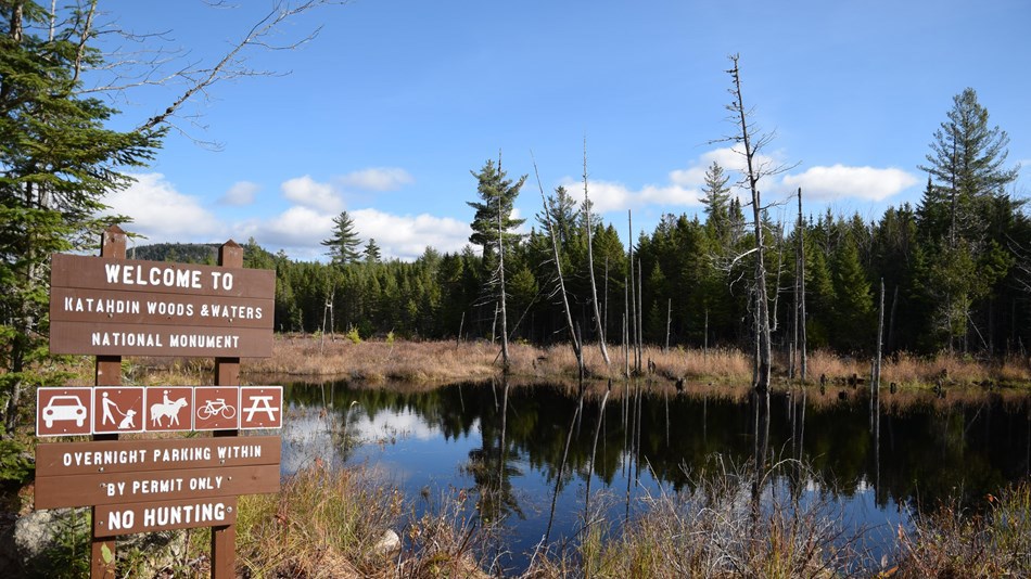 Katahdin Woods and Waters National Monument A wooden interpretive sign reading “Welcome to Katahdin Woods & Waters National Monument” is displayed near a still water pond.