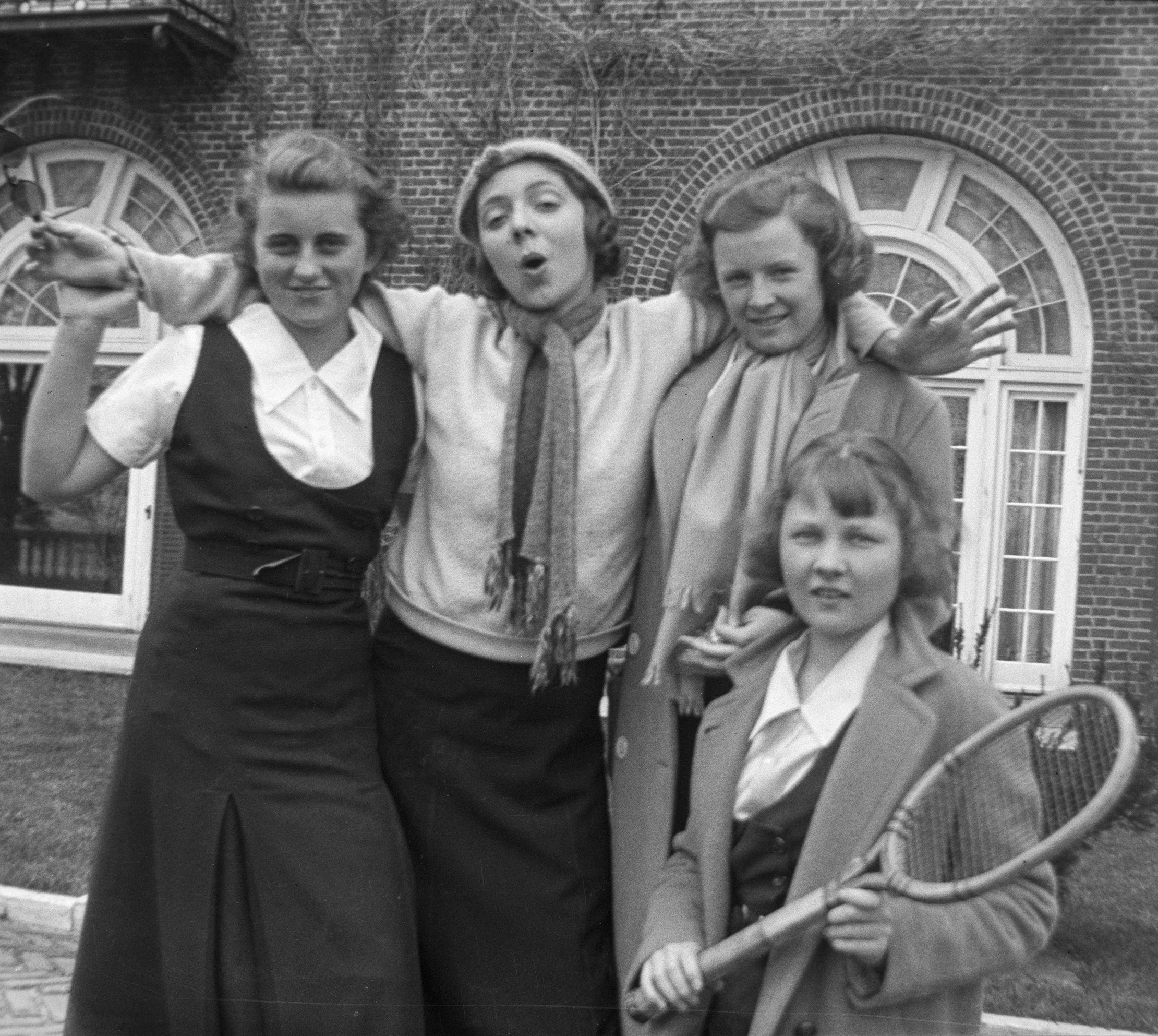 A black and white photo of four adolescent girls in skirts and curled hair posing in front of a brick building. One stands in center, her arms around the others’ shoulders, hand held by the girl left of her, and another is in front with a tennis racket.