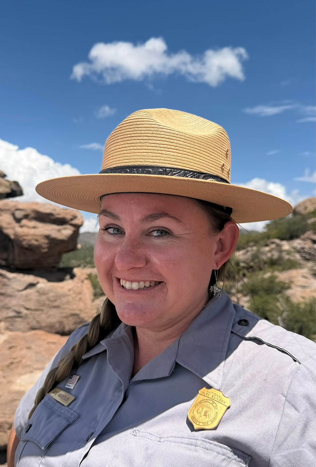A uniformed person smiles under a straw, flat-brimmed hat in a rocky landscape.