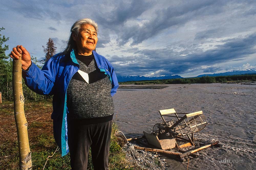 An Alaska Native woman stands in front of a muddy river.