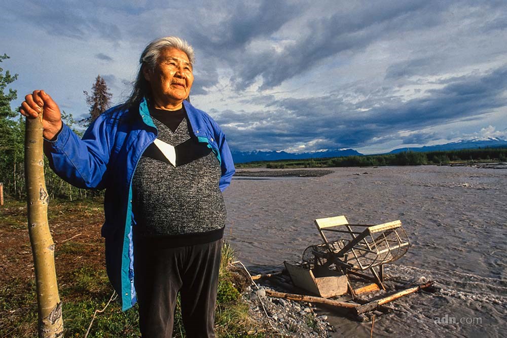 Katie-John-1994 WM_web An Alaska Native woman stands in front of a muddy river.