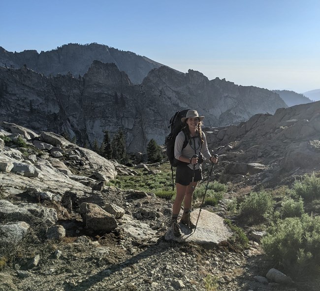 Woman wearing a backpack and hiking clothes stands in rocky setting with rugged peaks behind her.