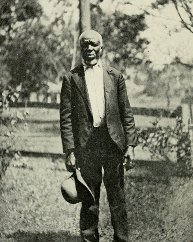 Kazoola A Black man stands in a suit outside holding his hat in his hand.