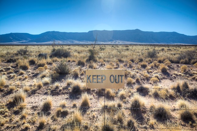 Faded sign reading "KEEP OUT" hanging on barbed wire fence at Trinity Test Site. Background shows mountain and a clear blue sky above the horizon line. Foreground and area below the horizon line show desert landscape with scrub vegetation.