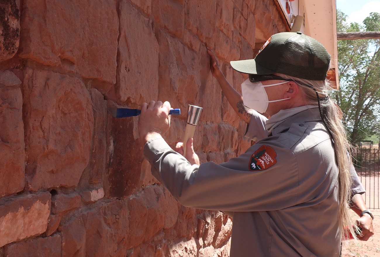 A man in National Park Service uniform, hat, and N95 mask uses a hammer and chisel to take a mortar sample from an old brick building.