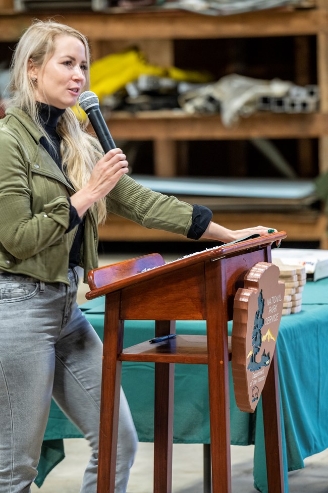 Woman in jeans and a green jacket stands at a lectern and speaks into a stick microphone Woman in jeans and a green jacket stands at a lectern and speaks into a stick microphone