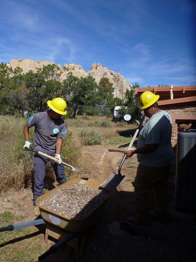 Two interns use shovels to move gravel into a wheelbarrow.