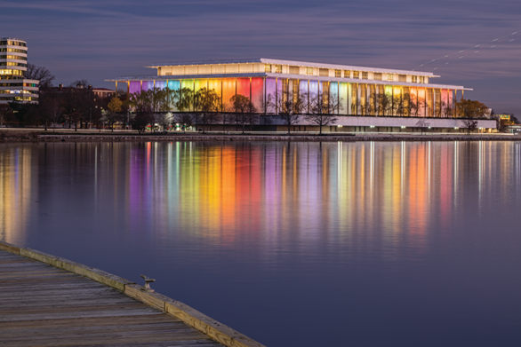 Kennedy Center Lighting Color HABS photograph at dusk showing the exterior of the Kennedy Center lit with rainbow colored lights. A reflection of the colorful structure can be seen in the Potomac River.