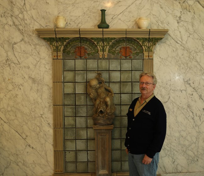 A middle-aged white man with white hair poses in an NPS volunteer uniform in front of an indoor fountain.