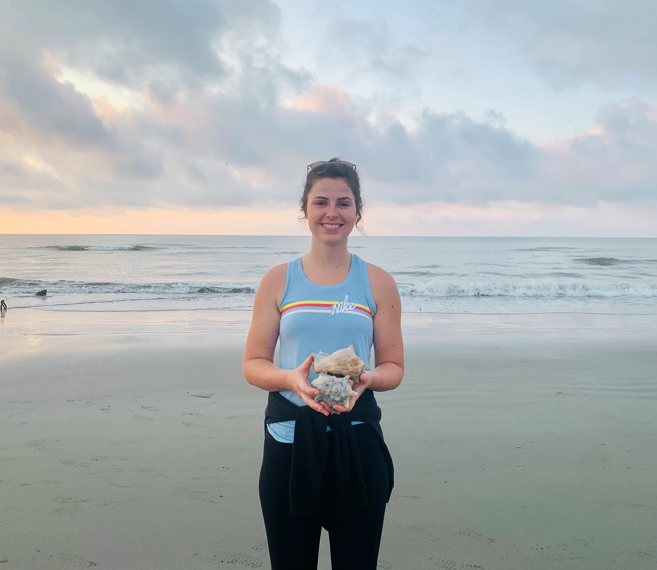 SC7 Woman stands holding two large seashells on sand in front of ocean, below sky in dimming sunset light