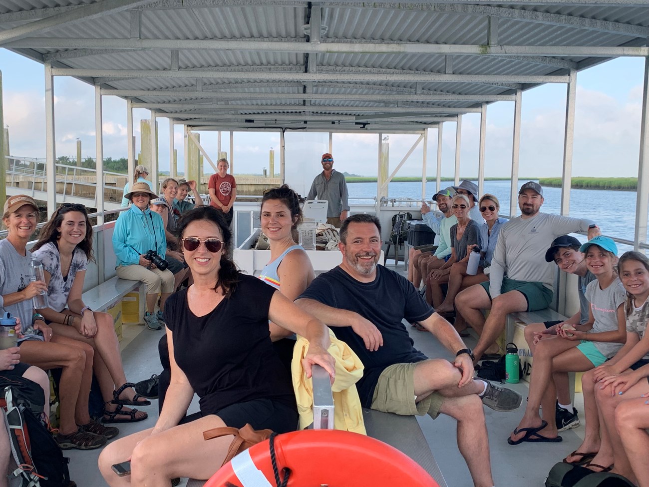 People seated on a small ferry boat pose for group photo. Ferry is moving across water below bright sky with a bit of marshland to right in background.