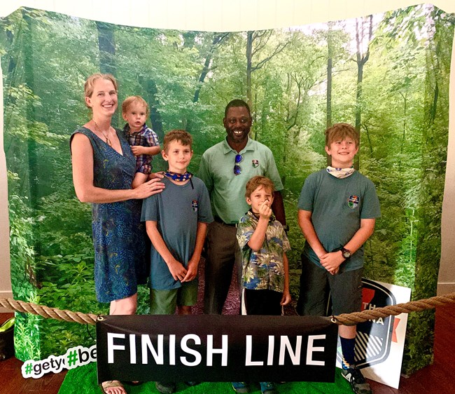 Six people (two adults and four children including one baby held in mother's arms) stand smiling in front of draped backdrop with faux green forest view and sign that reads "Finish Line"