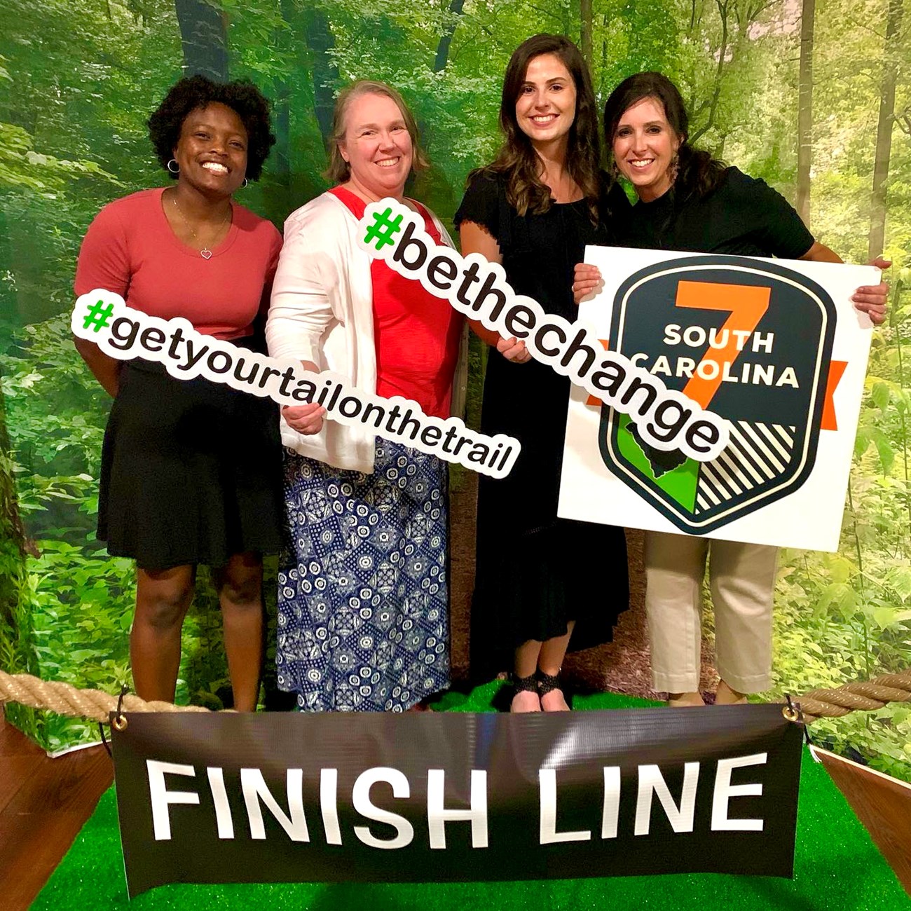 Four women stand in front of faux forest backdrop smiling, in front of sign reading "Finish Line". One (far right) holds sign reading "SC7". Two others hold signs reading: "#GetYourTailOnTheTrail" and "BeTheChange", respectively