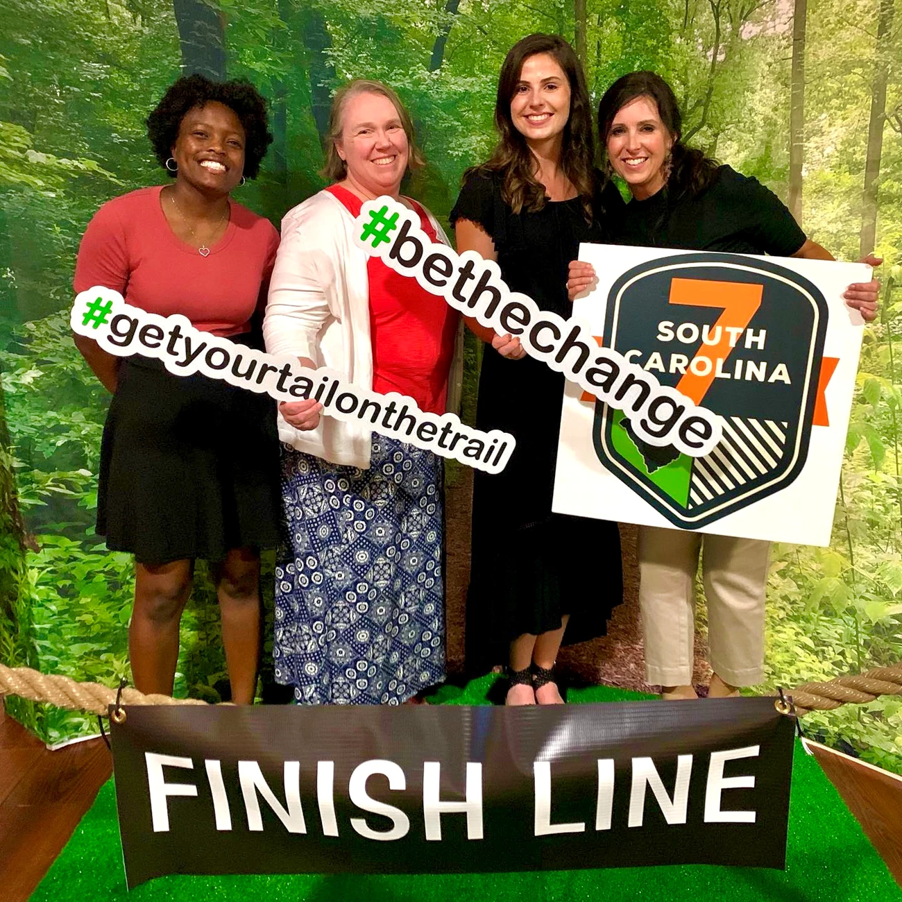 SC7 Four women stand in front of faux forest backdrop smiling, in front of sign reading "Finish Line". One (far right) holds sign reading "SC7". Two others hold signs reading: "#GetYourTailOnTheTrail" and "BeTheChange", respectively